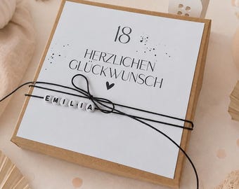 Könnte beinhalten: Eine quadratische Geschenkbox aus Karton mit einem weißen Etikett, das den Text 18 HERZLICHEN GLÜCKWUNSCH und ein kleines Herz zeigt. Die Box ist mit einer schwarzen Schnur umwickelt, die mit weißen Buchstabenperlen den Namen EMILIA bildet.