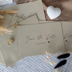 Könnte beinhalten: Beige "Save the Date" Karten mit eleganter Schrift, dem Datum 06.06.2026 und Herzdetails. Goldene Büroklammern und eine weiße Herzdekoration sind ebenfalls vorhanden, zusammen mit getrockneten Blumenelementen.