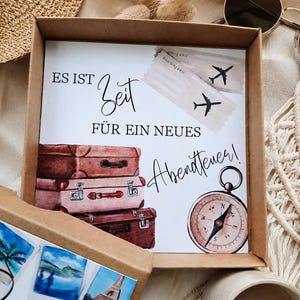 K&ouml;nnte beinhalten: Eine quadratische Geschenkbox aus Karton mit einer Karte, die den deutschen Text ES IST Zeit F&Uuml;R EIN NEUES Abenteuer! tr&auml;gt, illustriert mit drei gestapelten Vintage-Koffern, zwei Flugtickets und einem runden magnetischen Kompass.