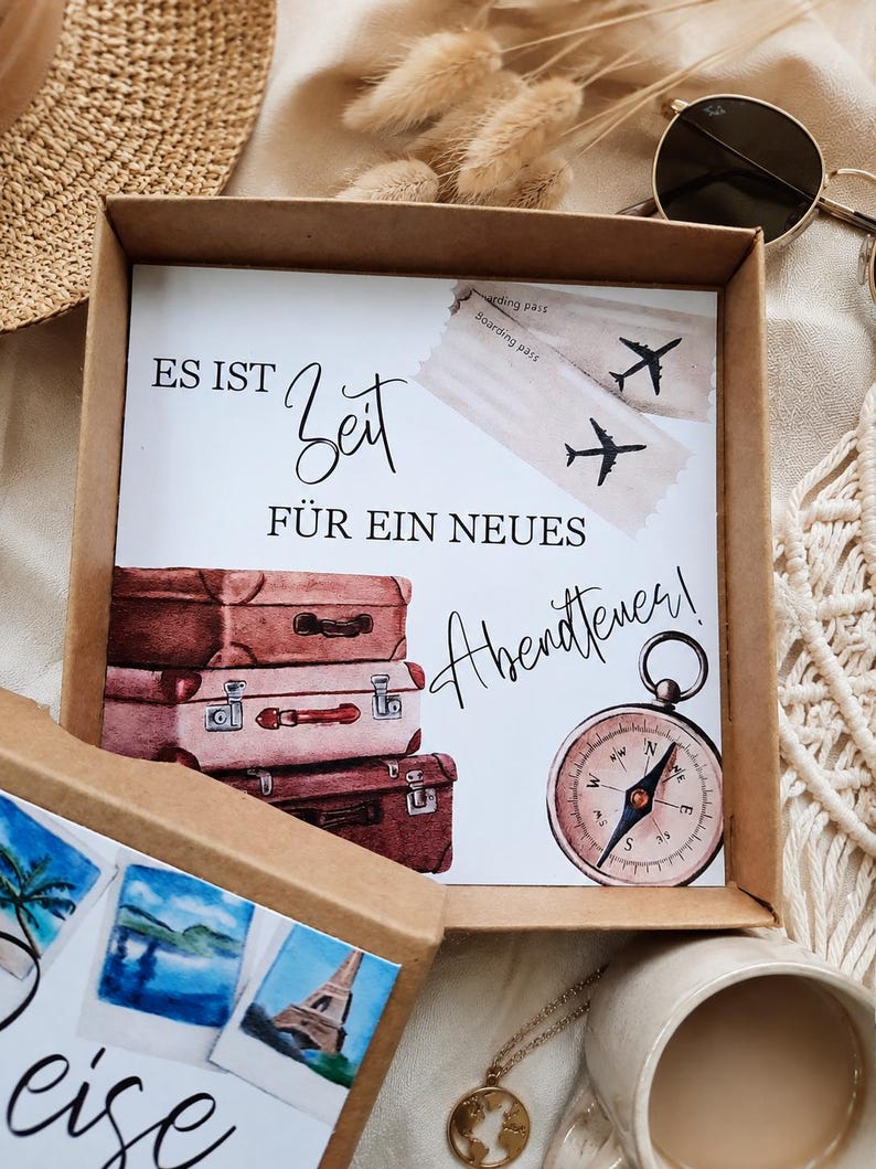 K&ouml;nnte beinhalten: Eine quadratische Geschenkbox aus Karton mit einer Karte, die den deutschen Text ES IST Zeit F&Uuml;R EIN NEUES Abenteuer! tr&auml;gt, illustriert mit drei gestapelten Vintage-Koffern, zwei Flugtickets und einem runden magnetischen Kompass.