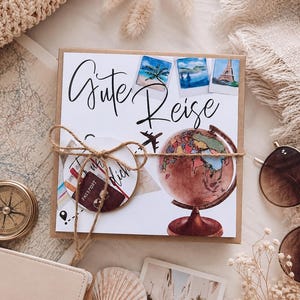 K&ouml;nnte beinhalten: Eine quadratische Geschenkbox mit einem wei&szlig;en Etikett, das den Text Gute Reise, einen Aquarell-Globus, ein kleines Flugzeugsymbol und drei Reisefotos zeigt. Die Box ist mit braunem Bindfaden verschn&uuml;rt und enth&auml;lt ein rundes Etikett mit Reisepass.