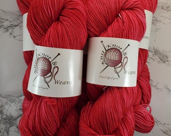 Puede incluir: Varios ovillos de hilo rojo teñido a mano. Cada ovillo está envuelto con una etiqueta blanca que dice "Knot & Weave Hand dyed yarn" con un logotipo de una bola de hilo y agujas de tejer. El hilo es de color rojo vibrante.