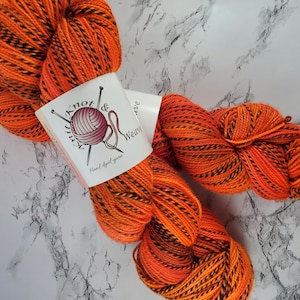May include: Orange and black hand-dyed yarn skeins with a label that reads "Knit & Weave". The yarn is a vibrant orange with black stripes, perfect for knitting or crocheting projects. The label features a ball of yarn and knitting needles.
