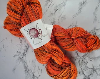 May include: Orange and black hand-dyed yarn skeins with a label that reads "Knit & Weave". The yarn is a vibrant orange with black stripes, perfect for knitting or crocheting projects. The label features a ball of yarn and knitting needles.