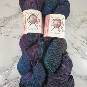 May include: Two skeins of hand-dyed yarn in shades of purple, blue, and green. Each skein has a label that reads "Knot Knit Wear" and "Blueberry Patch." The yarn is tightly wound and appears soft, ideal for knitting or crocheting projects.