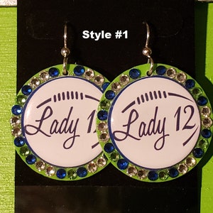May include: A pair of green and white round earrings with rhinestones. The earrings feature the text "Lady 1" and "Lady 12" in a cursive font.