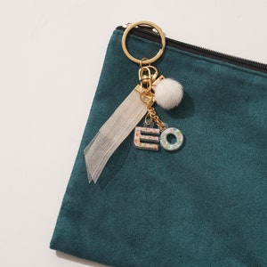 May include: A teal blue zippered pouch with a gold key chain attached. The keychain has a white pom pom, a tassel, and two charms that spell "EO".