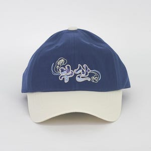 May include: A navy blue baseball cap with a white peak. The cap has an embroidered design in light blue and white on the crown. The cap has a white button on top.