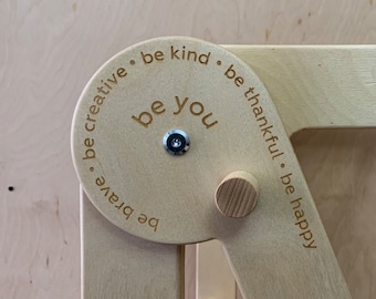 以下が含まれることがあります： 中央に「be you」の文字があり、周囲に「be creative, be brave, be kind, be thankful, be happy」と薄茶色のフォントで書かれた、木製のヒンジ付き構造。 この構造は明るい色の木材でできており、明るい色の木材の表面に取り付けられています。
