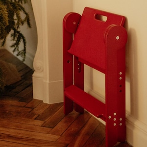 May include: A bright red, wooden step stool with a handle and two steps is leaning against a white wall. The stool is next to a Christmas tree and a white fireplace mantel. The floor is made of wooden planks.