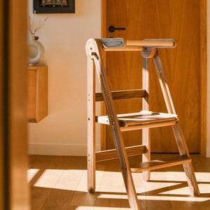 May include: A wooden step stool with a light brown finish. The stool has a ladder-like design with two steps and a top handle. Two framed artworks hang on the wall, and a wooden door is in the background.