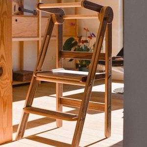 Könnte beinhalten: Ein Holztritt mit dunkelbraunem Finish. Der Hocker hat ein leiterartiges Design mit zwei Stufen und einem Griff oben. Der Hintergrund umfasst ein Holzregal und gerahmte Bilder.