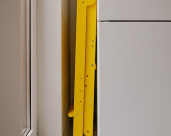 May include: A yellow folding step stool is tucked into a corner of a room. The stool is made of wood and has a simple design.
