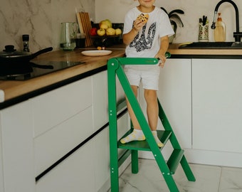May include: A young boy standing on a green step stool in a kitchen. He is wearing a white t-shirt with a black design and white socks. He is holding a cookie in his hand.