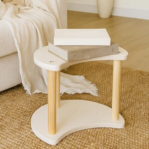 May include: A small, white, crescent-shaped side table with natural wood legs. Two neutral-toned books rest on the table's surface. The table sits on a woven jute rug, with a cream-colored sofa and a wicker chair in the background.