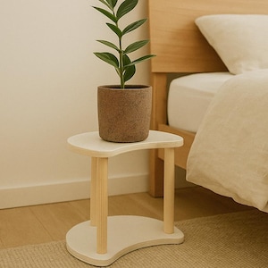 Puede incluir: Una pequeña mesa auxiliar de madera blanca con un diseño curvo y tres patas cilíndricas. Una planta en maceta con hojas verdes se encuentra encima. La maceta es marrón. El fondo incluye una cama con cabecero de madera y edredón beige.