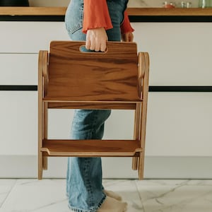 Pode incluir: Um banco de madeira com uma pega é segurado por uma pessoa. O banco é feito de madeira castanha clara e tem dois degraus. A pessoa está vestida com calças de ganga azuis e uma camisa laranja de manga comprida.