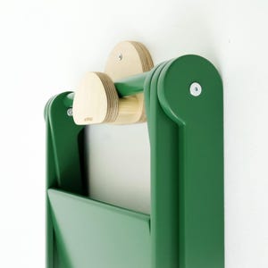 May include: A wall-mounted folding chair in a deep forest green finish, featuring light-colored natural wood accents on the top mounting bracket and hook. The chair is shown in a folded position against a plain white wall, secured with metal screws.