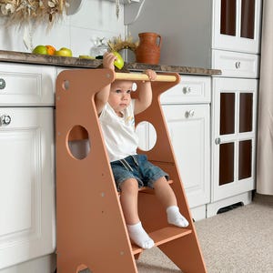 May include: A peach-coloured wooden kitchen step stool with a handle and two steps. The stool has a round cutout on the side. A child is sitting on the stool, holding onto the handle. The background includes white cabinets and a countertop.