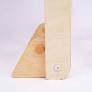 May include: Close-up of the wooden base of a light-coloured wooden chair. The base is made of two pieces of wood that are joined together with a screw. The wood has a natural finish.