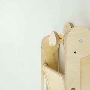 以下が含まれることがあります： 木目がはっきりと見える積層合板で作られた、明るい色の木製折りたたみ式ステップスツール。丸みを帯びたサイドパネル、中央の水平サポートバー、折りたたみ機能のための金属製ボルト機構を備えた構造。