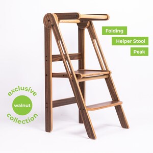 Puede incluir: Un taburete de ayuda plegable de madera con dos peldaños y un pasamanos. El taburete está hecho de madera de nogal y tiene un acabado natural. El taburete está etiquetado como "Folding Helper Stool Peak".