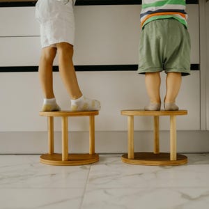 May include: Two small, round wooden stools with a light brown finish. The stools have a circular top and base, connected by four vertical legs. The stools are placed on a white and gray tiled floor. The stools are approximately 12 inches in height.