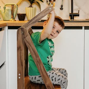 May include: A wooden step stool with a child sitting on it. The stool is brown and has a white label on the side that says "WOODIZZ". The child is wearing a green shirt with a dinosaur print and gray pants with a Batman print.