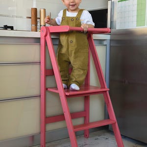 May include: A pink wooden step stool with a safety bar. The stool has two steps and is designed for children. A child wearing a white shirt and olive green overalls is standing on the top step.