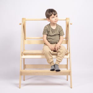 May include: A light-coloured wooden step stool with a child sitting on it. The stool has three steps and a safety bar. The child is wearing a green top, beige trousers, and grey socks. The background is white.