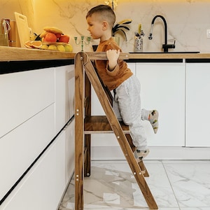 May include: A dark brown wooden step stool with two steps and a handrail. The step stool is in a kitchen with white cabinets and a marble countertop. A child is climbing the step stool. The step stool is designed to help children reach higher surfaces.