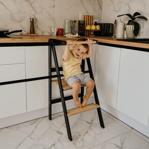 Puede incluir: Un niño se sienta en un taburete de paso de madera y negro con un pasamanos de madera. El taburete está en una cocina con armarios blancos y un salpicadero de mármol.