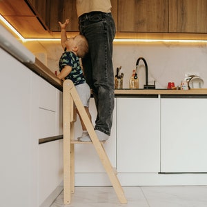 Puede incluir: Un niño pequeño de pie en un taburete de madera para alcanzar algo en una encimera de cocina. Un adulto está de pie detrás del niño, ayudándolo a alcanzar.