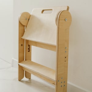 May include: A light wood step stool with a shelf and a handle on the top. The stool is made of plywood and has a simple design. The word "archi" is printed on the side of the stool.