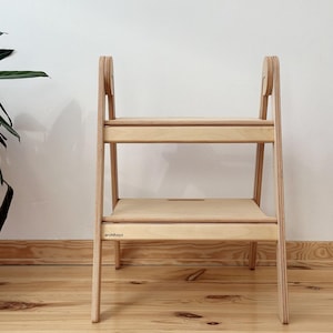 Puede incluir: Un taburete de madera de color claro con dos niveles y una estructura resistente. El objeto presenta un acabado de veta de madera natural, soportes laterales con detalles circulares y la marca architoyz impresa en el escalón inferior.