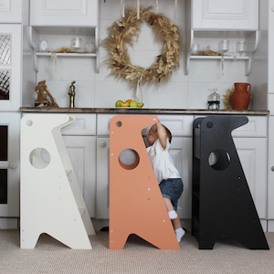 May include: Three wooden step stools in white, peach, and black. Each stool has a circular cutout and a unique angled design. A child is climbing on the peach-coloured stool. The stools are in a kitchen setting.