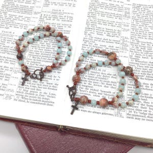 May include: Two beaded rosaries with brown and blue beads, a cross pendant, and a decorative clasp are displayed on an open book. The rosaries are resting on a page with printed text.
