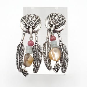 Pode incluir: Um par de brincos prateados com uma cabeça de águia esculpida no topo, duas penas prateadas pendentes e uma fileira central de contas. As contas incluem uma pedra rosa redonda, uma pedra azul-clara e uma pedra bege lisa e irregular.