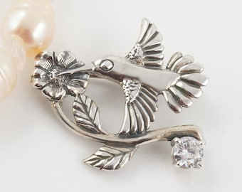 May include: A silver hummingbird pendant with a clear crystal on a pearl necklace. The hummingbird is perched on a branch with a flower.
