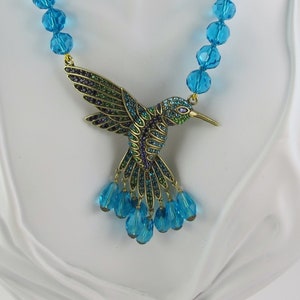 Könnte beinhalten: Eine türkisfarbene Perlenkette mit einem goldenen Kolibri-Anhänger. Der Kolibri ist mit grünen und blauen Kristallen verziert.