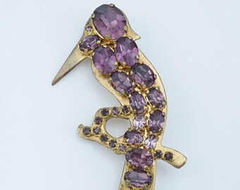 May include: A gold-toned bird brooch adorned with oval and round purple rhinestones. The brooch has a long, pointed beak and a curved body, with the gemstones arranged to mimic feathers. A vintage-style accessory.