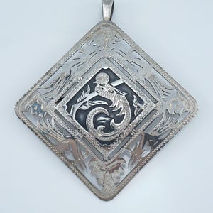 May include: A silver pendant with an ornate, openwork design. The pendant is diamond-shaped, with a central square featuring a detailed dragon motif. The outer frame has floral and geometric patterns. A small loop is at the top for a chain.