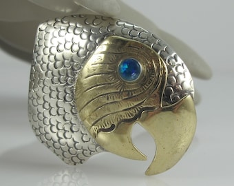 May include: A silver and gold parrot brooch with a blue gemstone eye. The parrot's head is detailed with textured feathers.