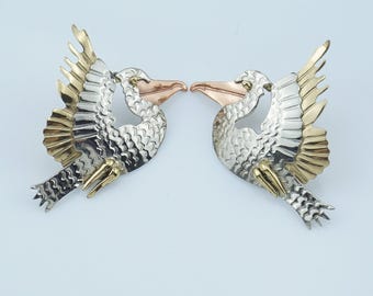 May include: Two bird-shaped brooches. Each brooch features a silver body, gold wings, and a copper-colored beak. The birds face each other, wings slightly spread, creating a symmetrical design. The background is a light blue.