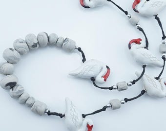 May include: A white swan necklace and bracelet set. The necklace features multiple white swan pendants with red beaks, strung on black cord with gray beads. The bracelet has matching gray beads. A unique jewelry set.