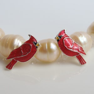 Op de afbeelding: Twee rode oorringen in de vorm van een kardinaalvogel met zilverkleurige randen en gele accenten. De oorbellen liggen op een streng grote, crèmekleurige parels. Een accessoire met een natuurthema.