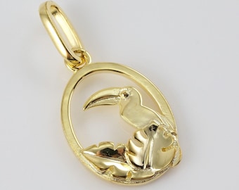 May include: A gold-colored pendant charm featuring a toucan bird perched on a leaf. The toucan is facing to the right and has a long, curved beak. The pendant is oval-shaped and has a loop at the top for attaching to a chain.