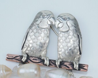 May include: A silver-toned brooch featuring two detailed parrot figures perched on a branch. The birds face each other, with intricate feather detailing. A strand of white pearls and clear stones is in the foreground.