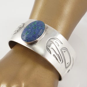 May include: Silver cuff bracelet with a blue and green stone set in the center. The bracelet is decorated with a repeating pattern of stylized animal faces.