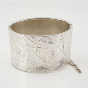 May include: A silver bangle bracelet with an engraved design of birds and bamboo. The bracelet has a chain attached to it.
